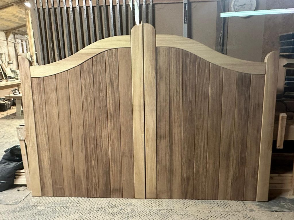 Bespoke iroko driveway gates in the finishing area before final treatment, showing vertical hardwood boards and curved top profile.