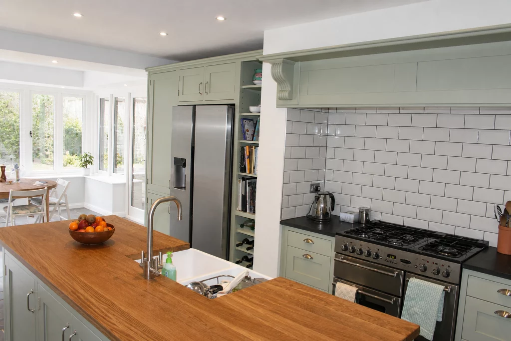 Cooking Apple Green Shaker kitchen with oak worktops and a tiled range recess, wide finished kitchen view with island, range and fridge housing.