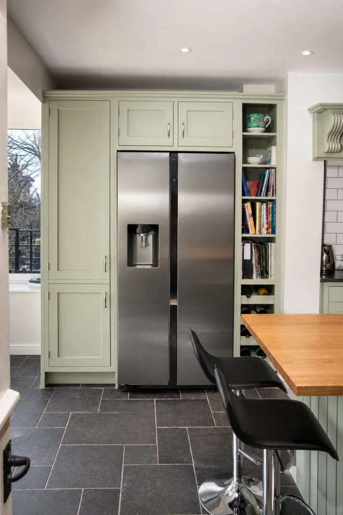 Cooking Apple Green Shaker kitchen with oak worktops and a tiled range recess, tall storage and fridge viewed from the end of the island.