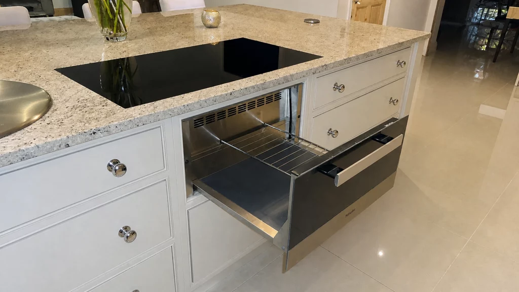 Dimpse-painted Shaker kitchen with a large granite island and integrated appliances, integrated warming drawer opened beneath the island hob.