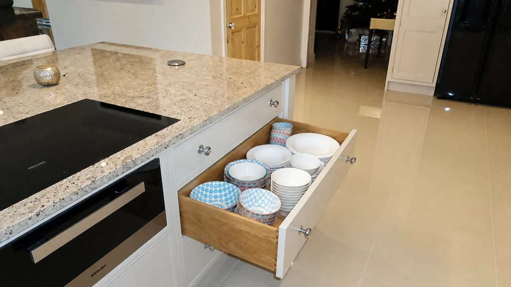 Dimpse-painted Shaker kitchen with a large granite island and integrated appliances, oak-lined crockery drawer opened beneath the worktop.