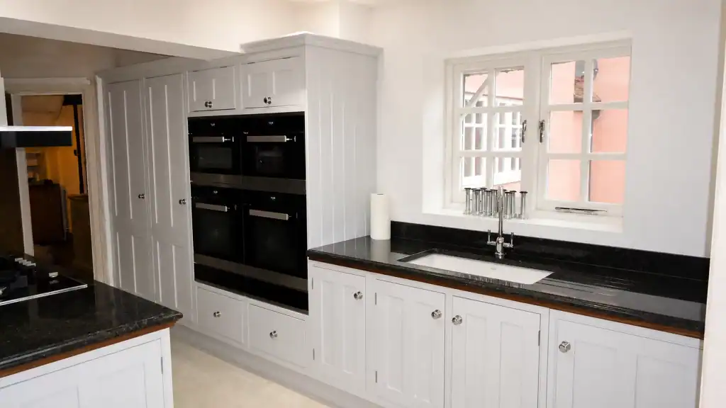 Painted pale-blue Shaker kitchen with dark granite worktops and oak details, sink run beneath the window with integrated black appliances.