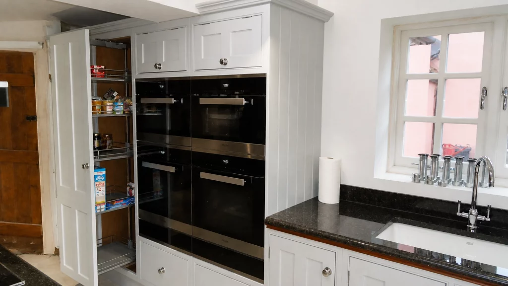 Painted pale-blue Shaker kitchen with dark granite worktops and oak details, tall pull-out larder beside the oven bank.