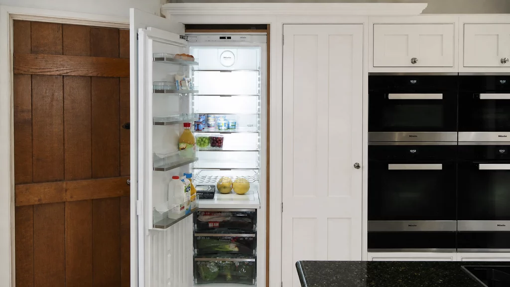 Painted pale-blue Shaker kitchen with dark granite worktops and oak details, open integrated refrigerator beside the oven bank.