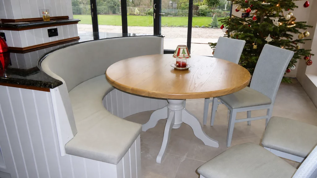 Painted pale-blue Shaker kitchen with dark granite worktops and oak details, round oak dining table framed by the half-round bench.
