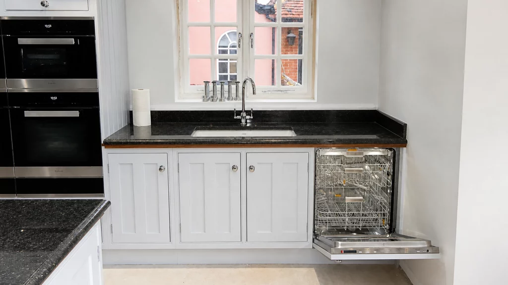 Painted pale-blue Shaker kitchen with dark granite worktops and oak details, sink cabinetry with integrated dishwasher open.